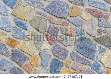 Its close up view of bright colorful stone wall. It's photo of multicolor stonewall. It view of blue, red, brown, grey and yellow srones. it's view of sidewalk. It's view of mosaic color stones
