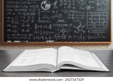An open textbook lies on a wooden desk in the foreground with a blurred background featuring a blackboard covered in various mathematical equations and diagrams.