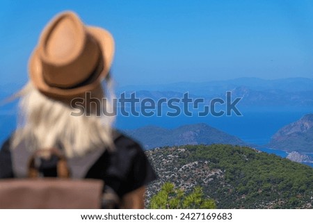 Beautiful view of the sea and mountains on a sunny summer day, in the foreground is out of focus a young female tourist in a hat with a backpack. Concept of travel, tourism.