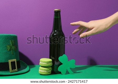 Glass bottle, green hat, macaroons, hand and clover leaf on purple background