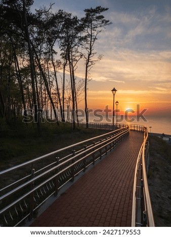 A tranquil pathway by the coast at sunset, featuring street lamps, trees, and a picturesque seascape. Royalty-Free Stock Photo #2427179553