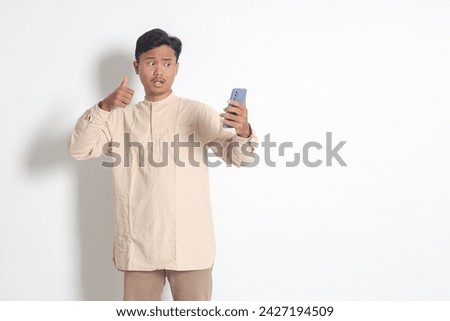 Portrait of young excited Asian muslim man in koko shirt holding mobile phone, taking picture of himself or selfie, saying hi and waving his hand. Social media concept. Isolated on white background