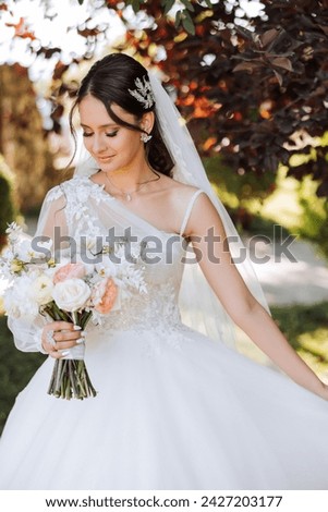 Wedding photo. A brunette bride in a lacy voluminous dress, holding a bouquet and walking in the garden, smiling sincerely. Gorgeous hair and makeup. Fashion and style. Celebration.
