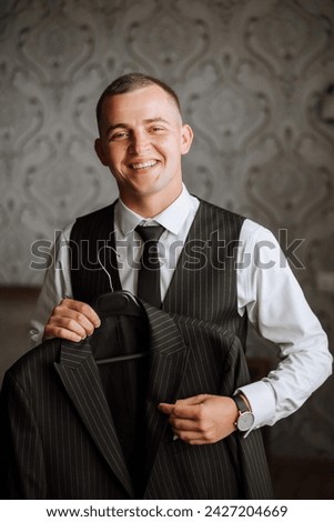 An elegant groom in a white shirt and a stylish tie is holding a jacket on a hanger. Details of the wedding morning in style. Royalty-Free Stock Photo #2427204669