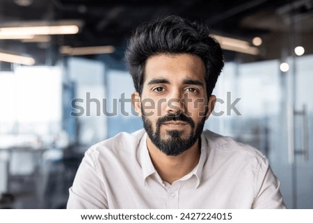 Close-up photo of a serious young Indian man with a beard and a shirt working in the office, sitting and looking confidently into the camera.