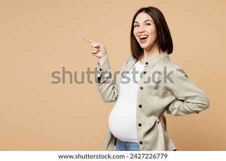 Side view young pregnant expectant woman future mom wear grey shirt with belly stomach tummy with baby point finger aside on area isolated on plain beige background. Maternity family pregnancy concept