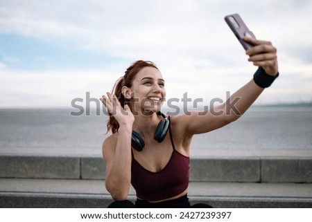 Joyful sports woman taking a selfie wearing headphones sitting outside. Young adult fit athletic female in sportswear taking a picture with smartphone