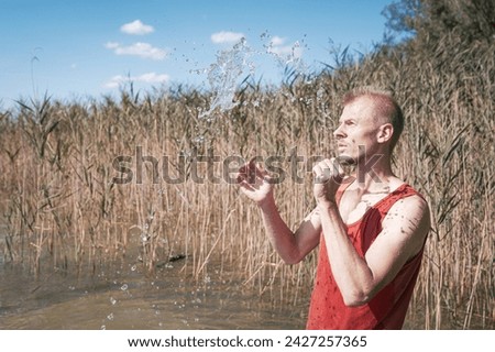 A man wearing a muscle shirt stands on the shore of a lake and has scooped water into the air. Royalty-Free Stock Photo #2427257365
