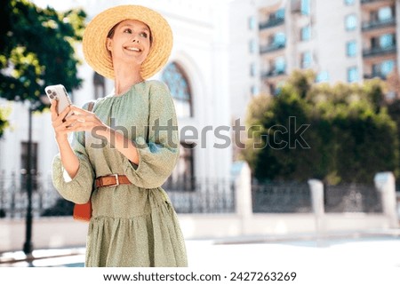 Young beautiful smiling woman in trendy summer sundress clothes. Carefree female posing in street at sunny day. Positive model holds smartphone, looks at cellphone screen, uses mobile apps. In hat Royalty-Free Stock Photo #2427263269