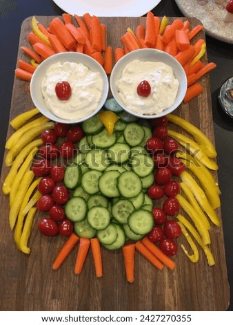 The picture shows an owl made of vegetables. The owl has a body made of cucumber slices, eyes made of cherry tomatoes, and feathers made of carrot sticks. There are two bowls of ranch dressing.