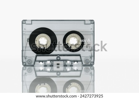 An audio cassette in a transparent case stands on a white background with reflection