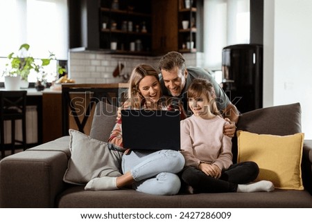 Joyful family of three spends quality time together on the living room sofa, sharing a moment around a laptop in their comfortable home Royalty-Free Stock Photo #2427286009