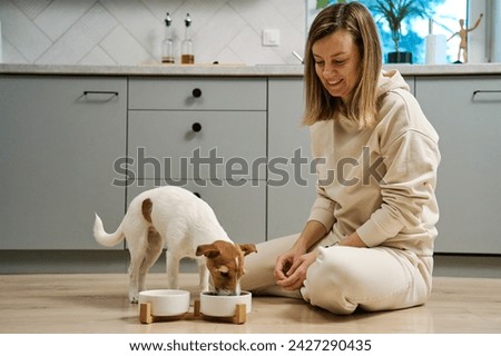 Woman putting food bowl with feed for her dog on the floor in kitchen, Female owner spending time together with pet at home, Animal feeding and pet care Royalty-Free Stock Photo #2427290435