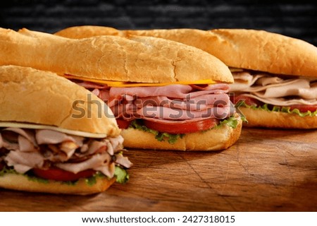 Assorted delicious baguette sandwiches filled with thinly sliced ham or salami and fresh green lettuce or basil arranged in an oblique row on an old wooden table .
