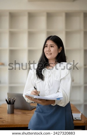 Female office worker holding documents and pen thinking about project and taking notes in office