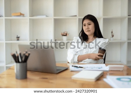 Young businesswoman takes a coffee break while working using a laptop in a bright office. Think and look away