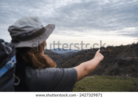 Back view of young traveler woman in hiker equipment points with finger at the mountains background, mountaineer enjoy beauty of the nature, focus on the hills, copy space