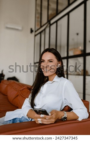 attractive smiling woman sitting on sofa at home cozy relaxed using smartphone wearing white t-shirt and jeans listening to music on earpods