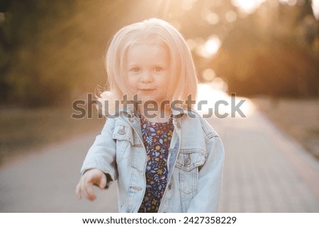 Cute kid girl 2-3 year old with blonde hair wearing denim jacket outdoor over dun light. Looking at camera. Childhood. 