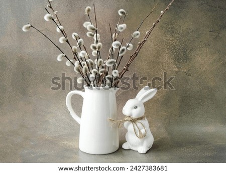  Easter decorative composition on a brown background, minimalistic composition, a white vase with willow branches and a white rabbit stands on the table.  Concept of decorated houses for Easter.