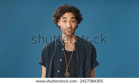 An attractive man with curly hair, dressed in blue shirt, listens and disagrees while looking at camera isolated on blue background in studio