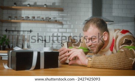 Blogger man taking picture or recording video on mobile phone of gift box on kitchen table. Smiling man using smartphone indoor at domestic kitchen
