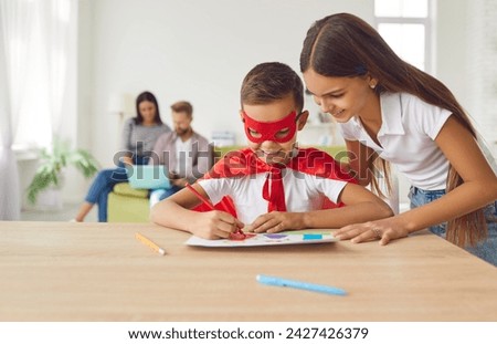 Happy little boy in superhero costume draws picture with help of his older sister. Boy in red superhero cape and glasses sits at table and draws at home against background of his parents.