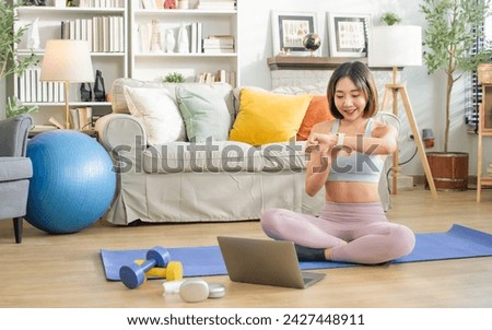 Asian Sportive beautiful woman smiling with happiness, looking at watch on wrist to check heart rate, calories burnt, sitting in living room at cozy home. Technology, Exercise, Healthy Concept