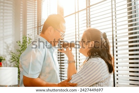 Asian adult couple wearing casual clothes, standing in cozy home, looking each other with warmth and love, smiling with happiness, giving anniversary cake. Relationship Concept