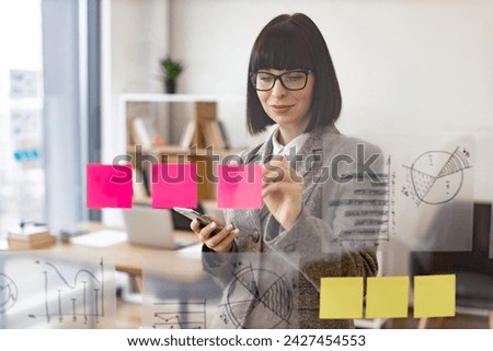 Portrait of beautiful young woman standing in front of glass board and typing on smartphone. Female office worker in glasses and stylish suit using digital phone in company.