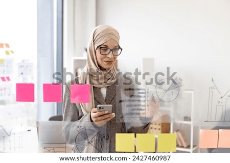Pensive arab businesswoman looking on phone while analyzing growth of charts on glass board at modern office. Concept of business strategy and digital marketing.