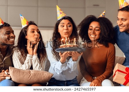 Cheerful group of multicultural friends gathering in living room to celebrate birthday, lady blowing candles on cake making her bday wish, wearing festive hats at home. Celebration and friendship