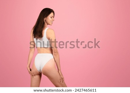 Side view of a content happy caucasian woman in a white sports bra and panties, with a subtle smile, standing confidently against a soft pink background, evoking wellness Royalty-Free Stock Photo #2427465611
