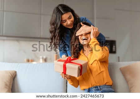 Joyful European lady surprises her girlfriend playfully covering eyes before giving gift box, celebrating special moment together in modern living room at home. Holiday Celebration, Valentine Day