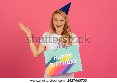 Joyful Bday Celebration. Smiling young blonde woman in party hat holding Happy Birthday bag, looking at camera, showing surprise present and raising arm in joy, on pink studio background