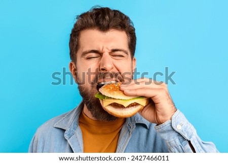 Funny bearded man enjoying cheat meal savoring burger against blue studio background, closeup portrait. Guy biting delicious fast food, tasting takeaway cheeseburger with appetite
