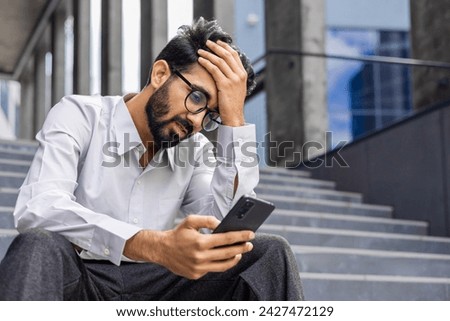 Young man depressed, sad dissatisfied and unhappy outside office building, holding phone, reading bad news from smartphone, businessman in shirt after work. Royalty-Free Stock Photo #2427472129