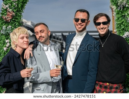 Newlywed gay male couple poses with friends for a memorial photo. They are enjoying a few glasses of white wine, outdoors, under a mistletoe.