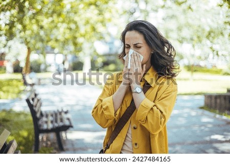 Close-up of a young woman blowing nose with tissue paper at the park. Woman with with allergy symptom blowing nose. Young pretty woman sneezing in front of blooming tree. Spring allergy concept
