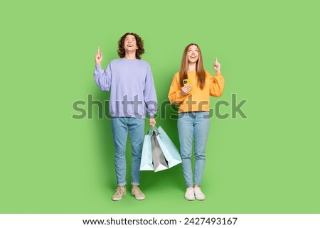 Full length portrait of two astonished people shop bags smart phone look indicate finger up empty space isolated on green color background