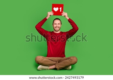 Full size photo good mood handsome guy wear red shirt sit holding red social media like over head isolated on green color background