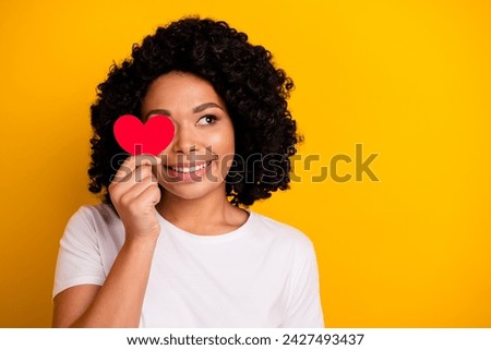 Photo of pretty good mood woman with perming coiffure hold red heart on eye look at promo empty space isolated on yellow color background