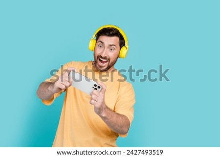 Photo of addicted gamer guy wearing wireless headphones with dolby technology sound playing mobile games isolated on cyan color background