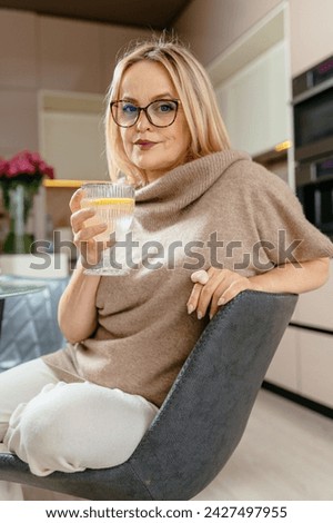 Vertical lifestyle portrait of 50s aged blond woman drinking water in eyeglasses home owner sitting in modern kitchen interior.