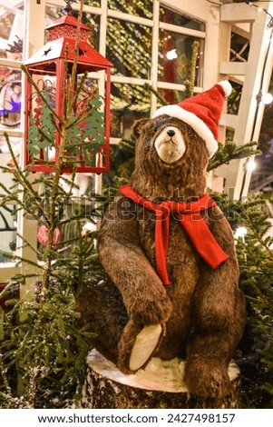 A bear figurine in a red hat and scarf sits on a stump among the Christmas trees. Christmas decoration.