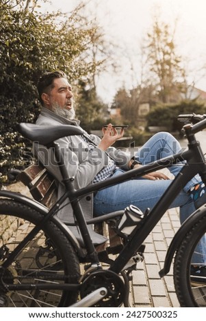 A middle-aged man with a beard is sends voice message while taking a break on a park bench, with his bicycle propped beside him. Royalty-Free Stock Photo #2427500325