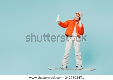 Full body young woman wear warm windbreaker jacket hat ski goggles snowboarding do selfie mobile cell phone mask travel rest spend weekend winter season in mountains isolated on plain blue background