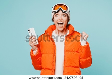 Skier winner young woman wearing warm padded windbreaker jacket hat ski goggles mask hold use mobile cell phone travel rest spend weekend winter season in mountains isolated on plain blue background