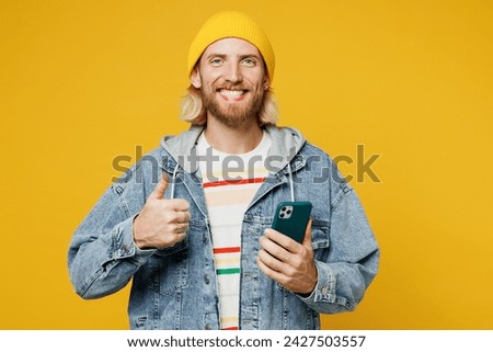 Young smiling blond man he wearing denim shirt hoody beanie hat casual clothes hold in hand use mobile cell phone show thumb up isolated on plain yellow background studio portrait. Lifestyle concept