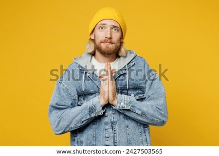 Young blond man he wears denim shirt hoody beanie hat casual clothes hold hands folded in prayer gesture, begging about something isolated on plain yellow background studio portrait. Lifestyle concept Royalty-Free Stock Photo #2427503565